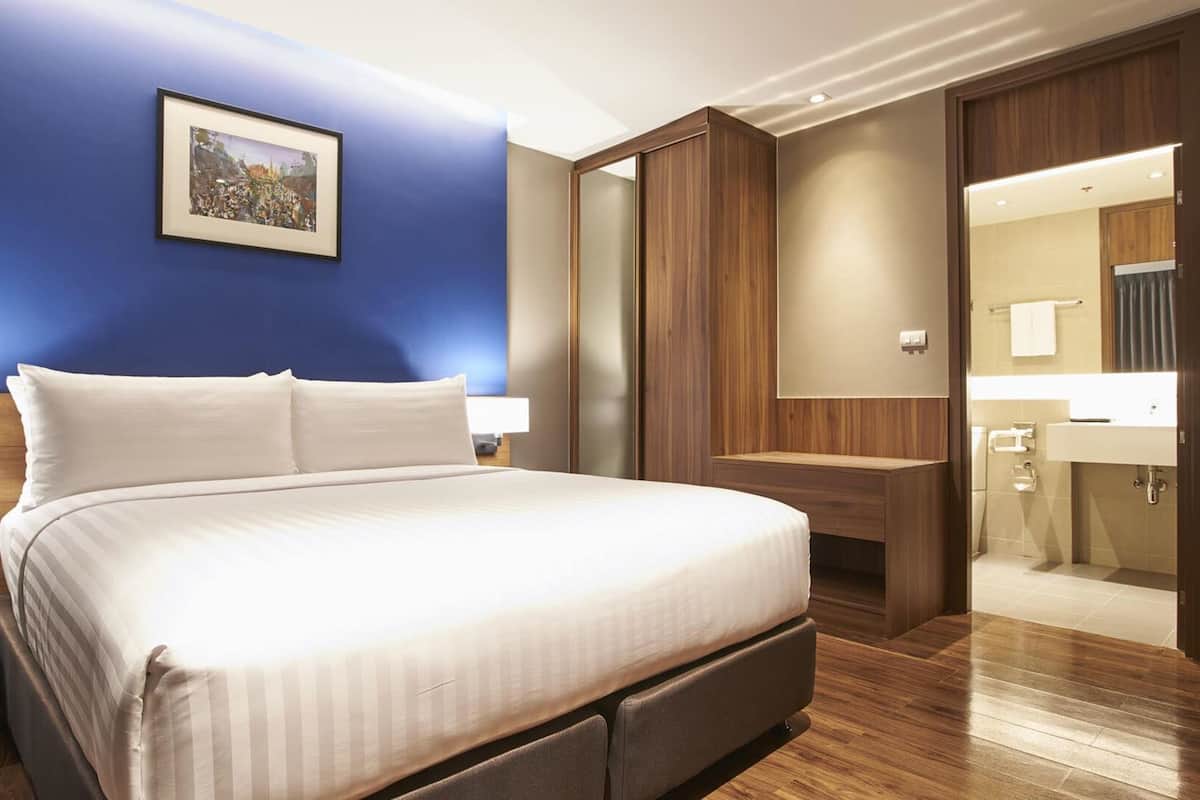 A modern hotel room with a large bed, bedside lamps, a wooden floor, a bathroom with a mirror and sink, and a framed picture on the wall.