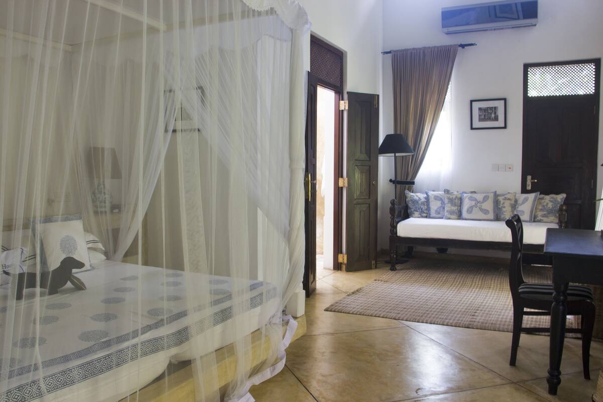 A bedroom with a bed covered in a mosquito net, a sofa with patterned cushions, a wooden table, and a chair.