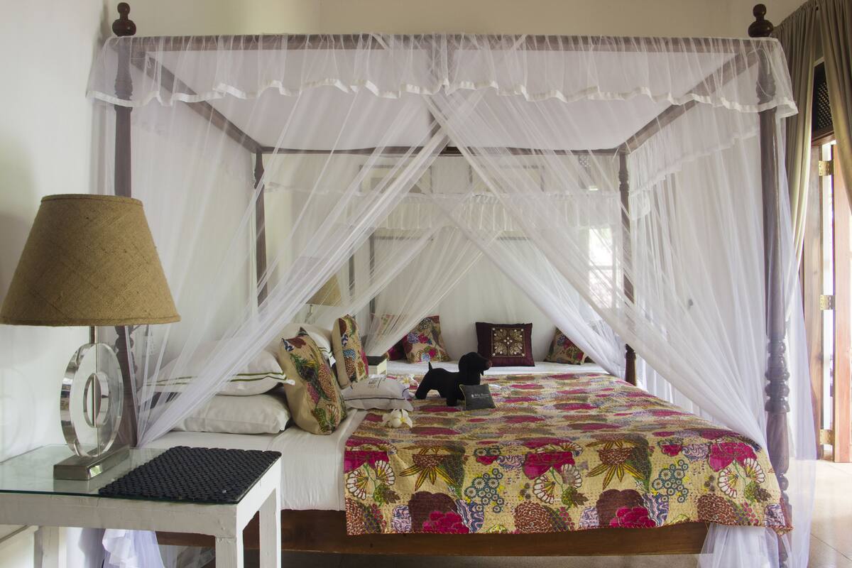 A four-poster canopy bed with a floral bedspread, a bedside table with a lamp, and a window with curtains.