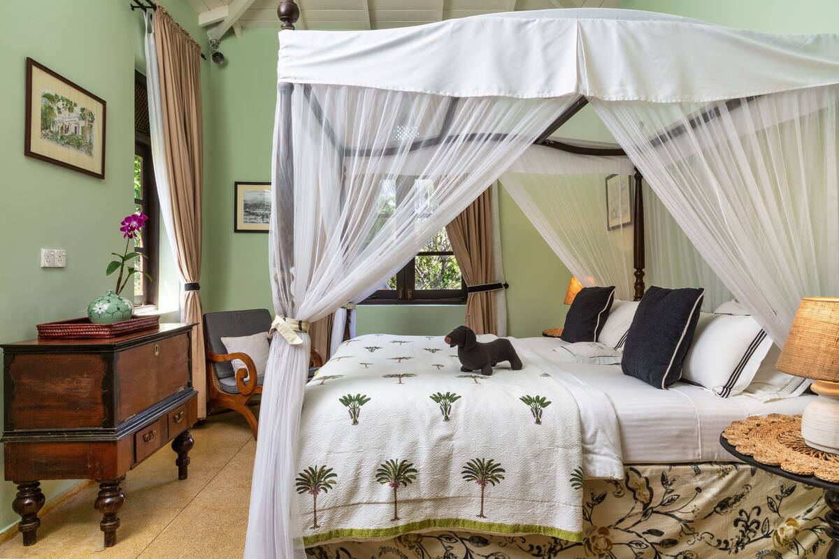 A four-poster canopy bed with a white bedspread featuring palm tree patterns, a wooden nightstand with a vase, and a chair with a cushion.