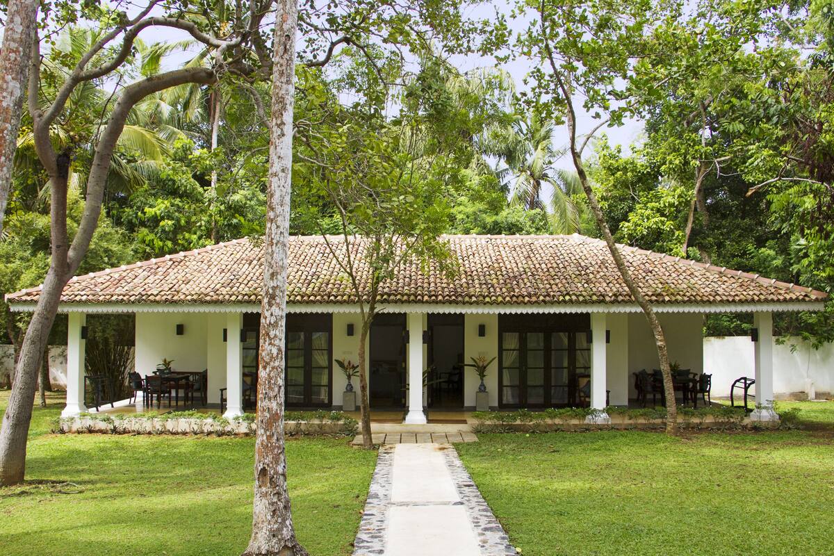 A two-story house with a tiled roof, a covered porch with chairs, and a pathway leading to the entrance.