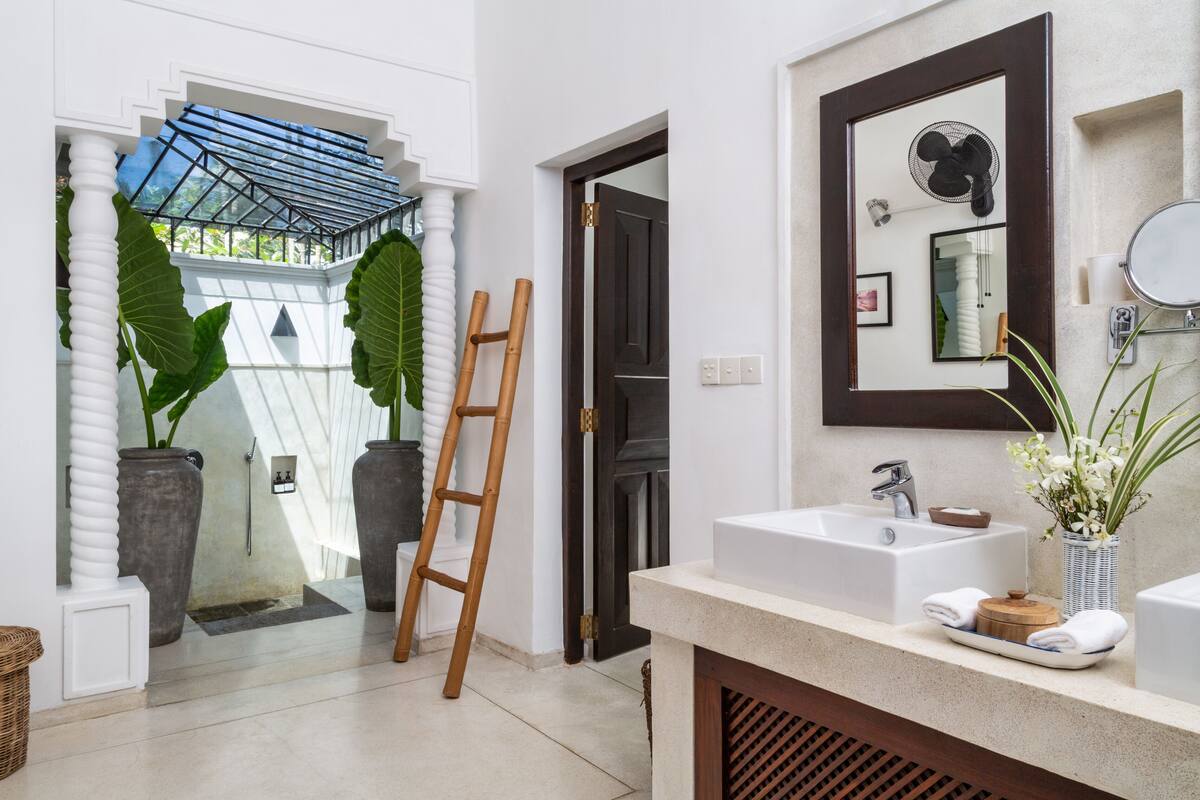 A bathroom with a large mirror, a sink, and a fan. There is a ladder leaning against the wall and a potted plant on the counter.