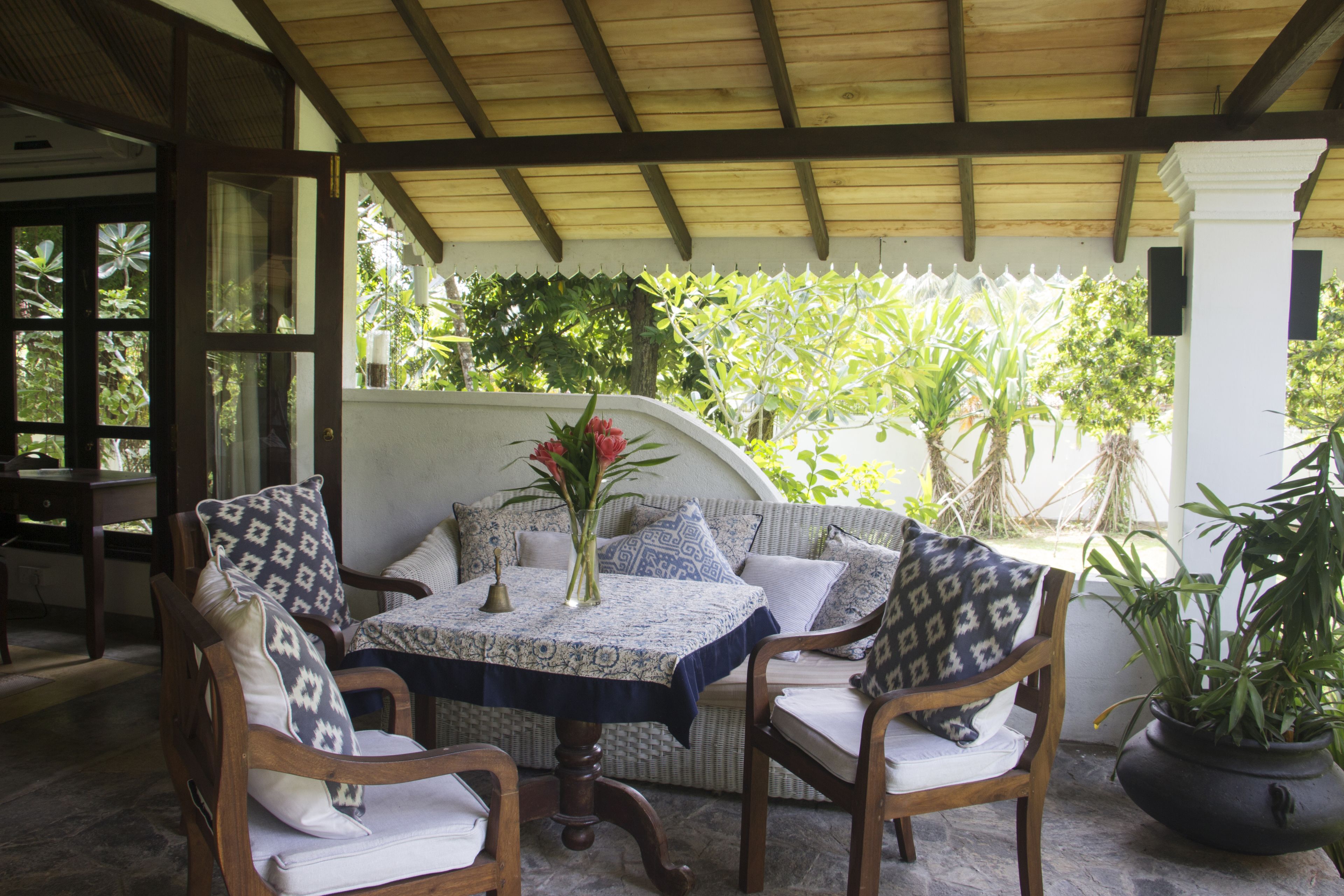 A covered patio with a table and chairs, surrounded by potted plants and a view of greenery.
