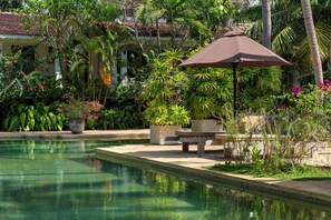 Un área de piscina con un banco y una sombrilla, rodeada de exuberante follaje tropical y un edificio con techo de tejas.