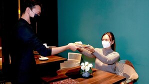 Una persona sirve comida a otra en una mesa de madera con un jarrón de flores blancas.