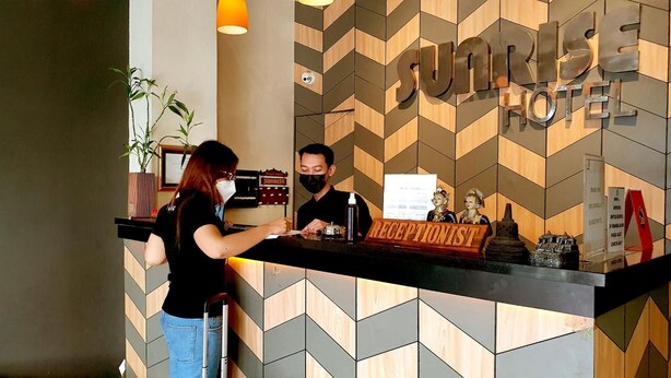 Mostrador de recepción en el Hotel Sunrise con un diseño moderno de pared geométrica, una recepcionista y un huésped.