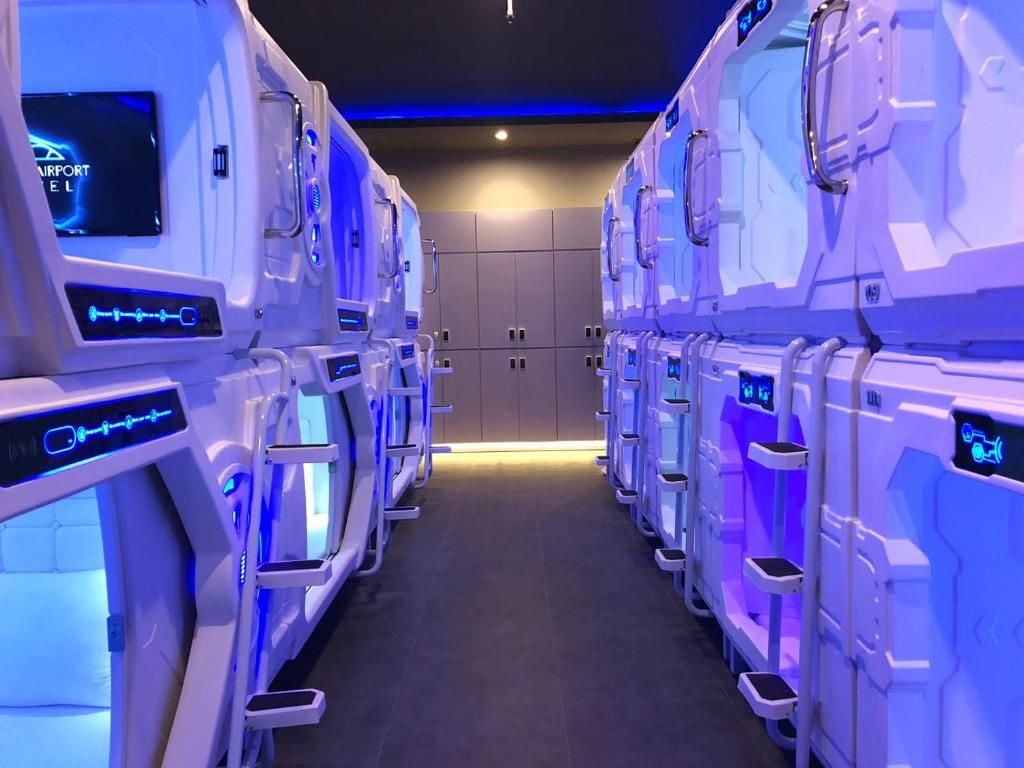 A row of futuristic-looking massage chairs with control panels, arranged in a corridor with a dark floor and walls.