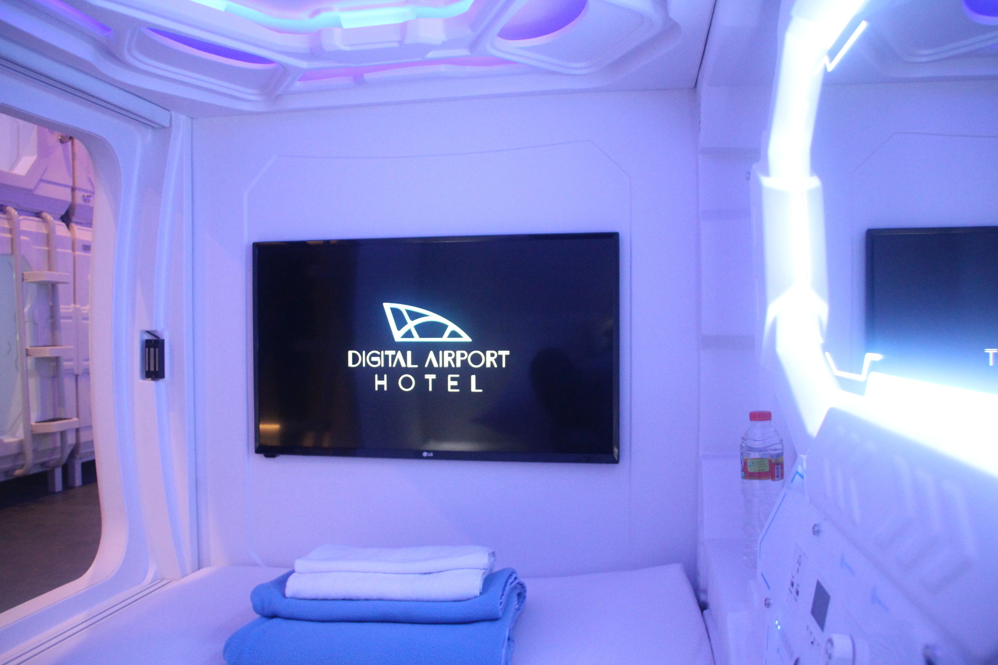 Una habitación con una cama, un televisor en la pared que muestra 'Digital Airport Hotel' y una botella en la mesita de noche.