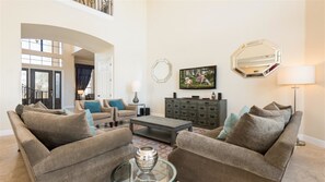A spacious living room with a high ceiling, a large window, and a view of the outdoors. There is a sofa set, a coffee table, and a TV mounted on the wall.