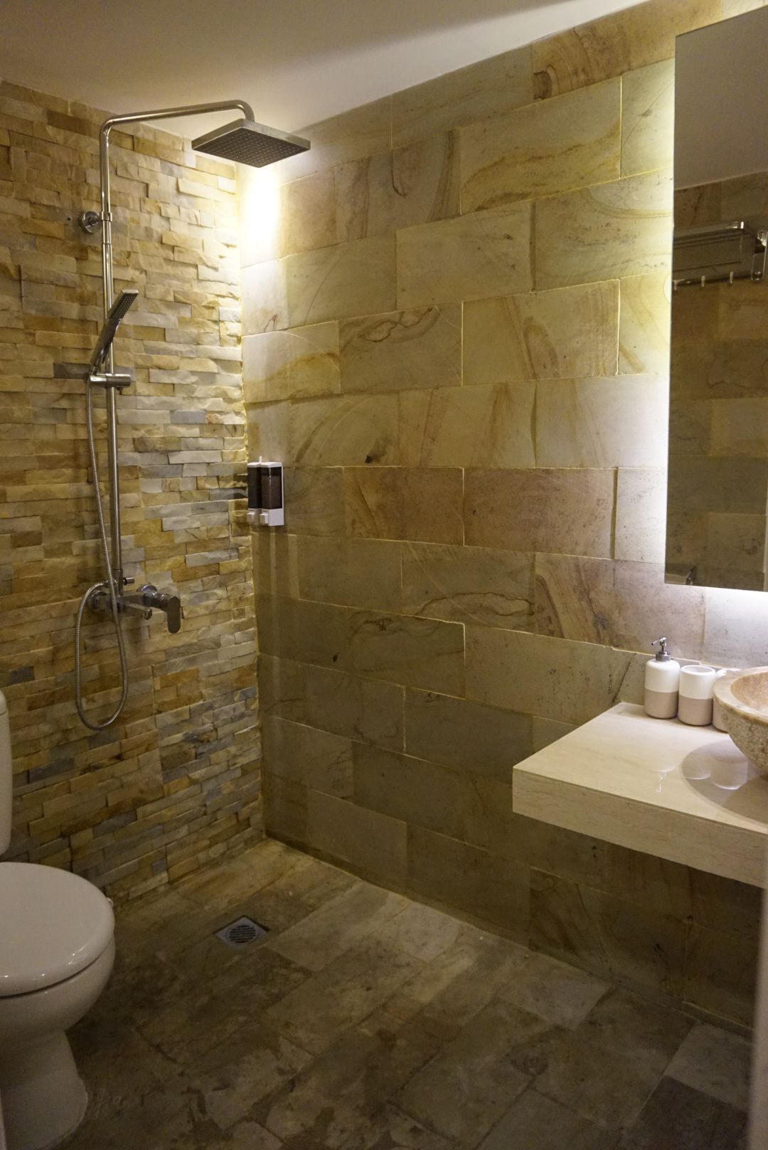 A bathroom with a stone wall, a shower, a toilet, and a sink with toiletries.
