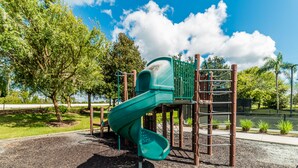 Ein Spielplatz mit einer grünen Rutsche, umgeben von Bäumen und blauem Himmel.