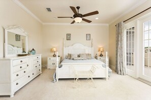 Quarto com cama branca, ventoinha no teto, abajures, cômoda e uma janela grande com cortinas.