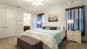 A bedroom with a bed, nightstands, a bench, a ceiling fan, a window with curtains, and a painting on the wall.