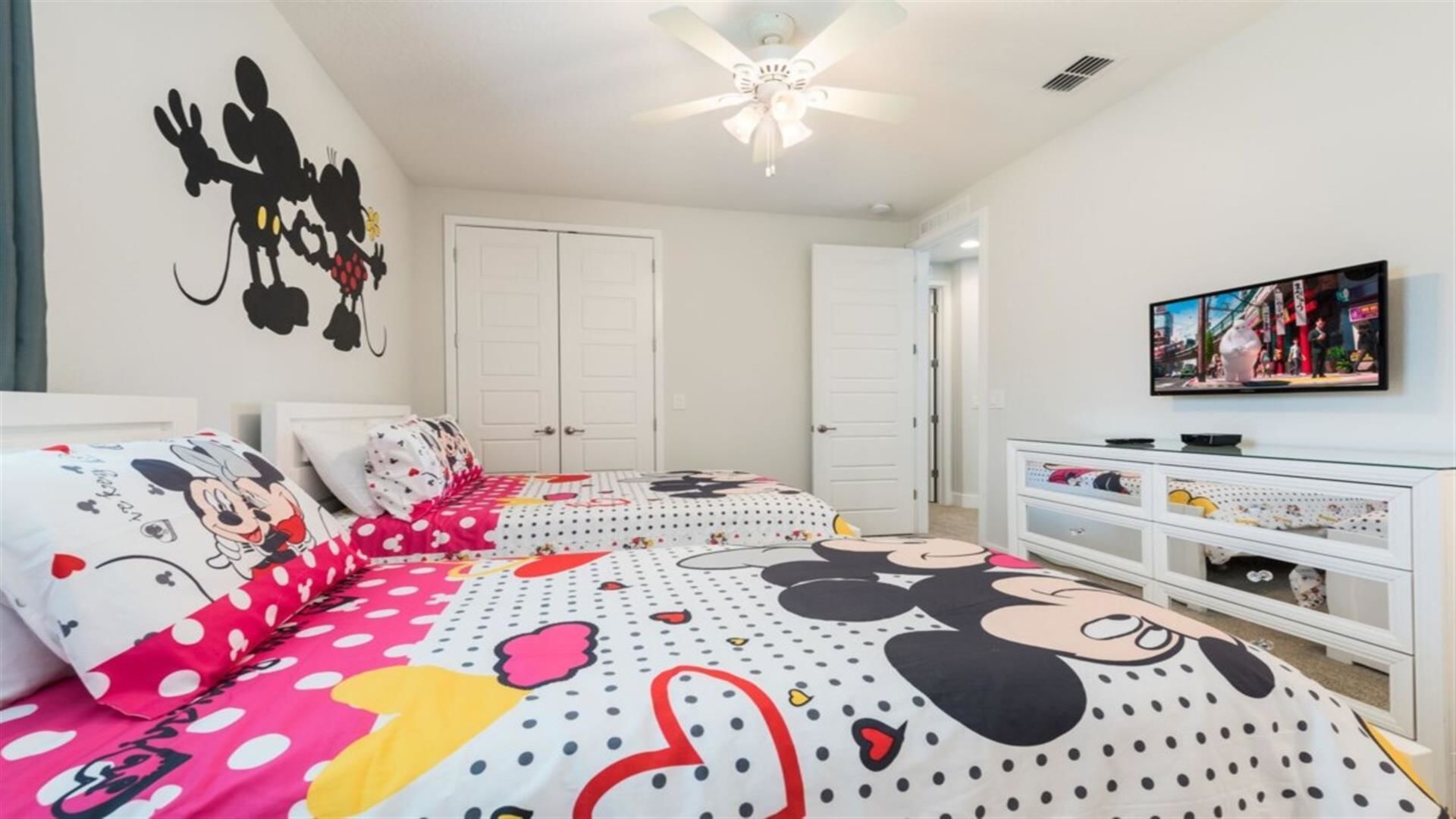 A bedroom with two beds, each with Mickey Mouse themed bedding, a ceiling fan, a TV on the wall, and a white dresser.