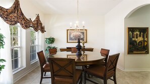 A dining room with a round table, six chairs, a chandelier, and a painting on the wall.