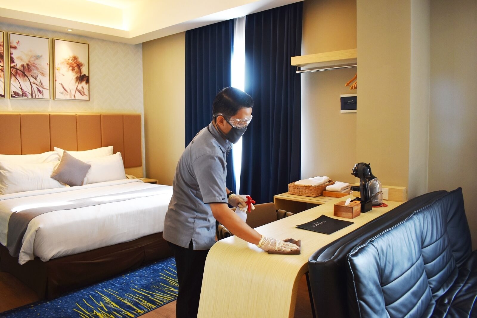 Habitación de hotel con cama, escritorio y silla. Se ve a una persona con mascarilla y guantes, posiblemente un empleado del hotel, de pie cerca del escritorio.