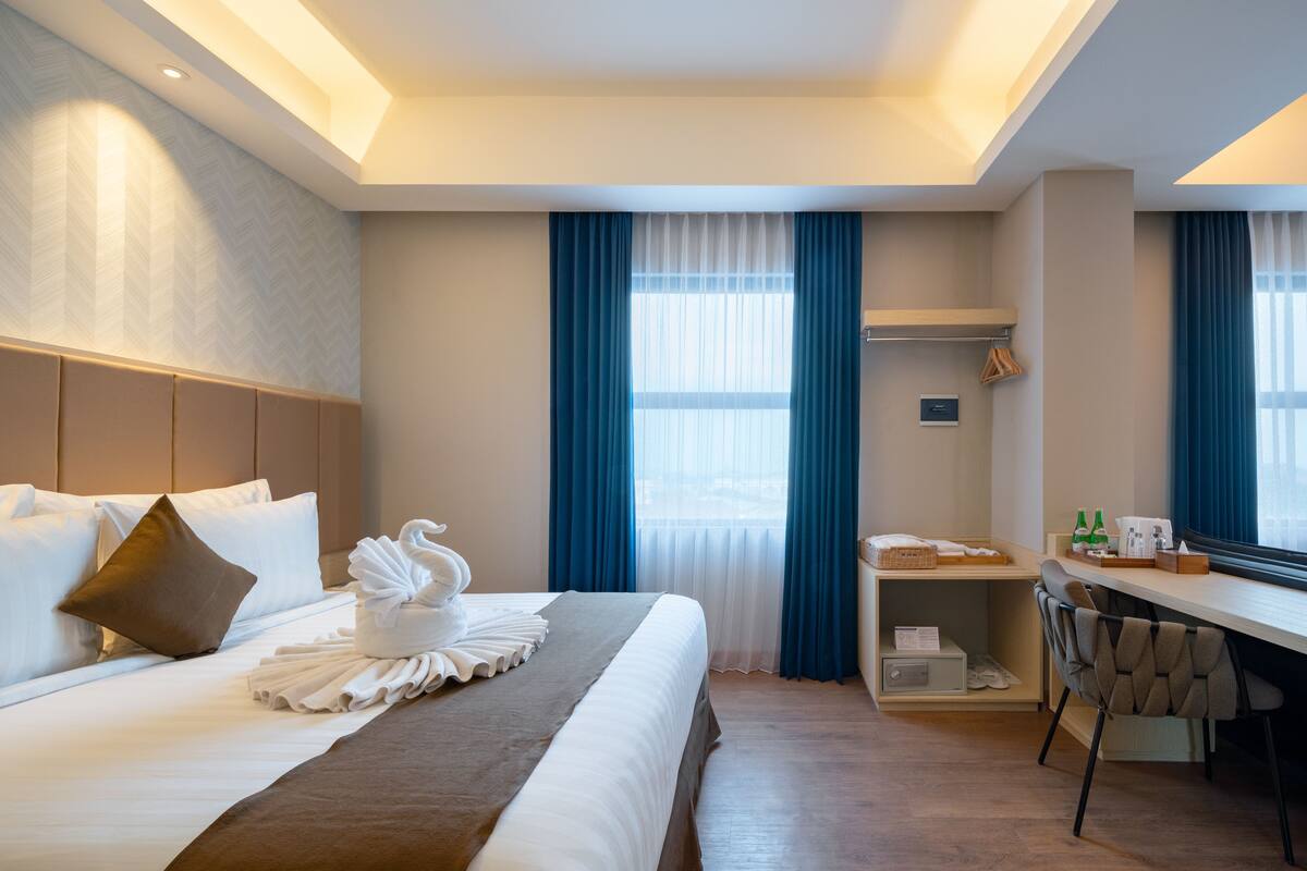 A hotel room with a neatly made bed, a desk with a chair, a shelf with personal items, and a window with curtains.