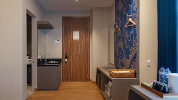 Habitación de hotel con puerta de madera, kitchenette, baño con espejo y toallero, y mostrador con cesta y botellas de agua.