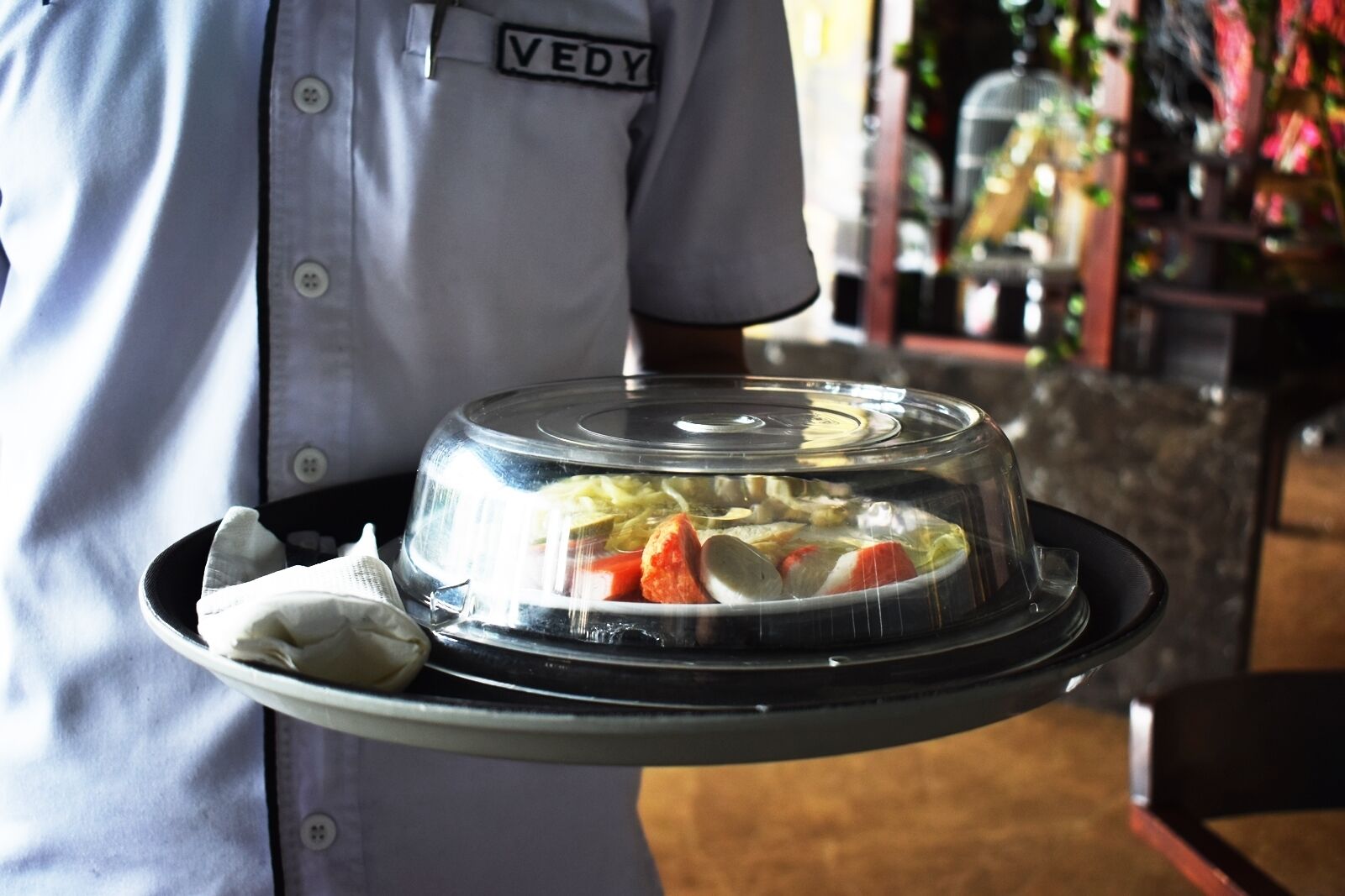 A person holding a covered food tray with a name tag reading "VEDY".