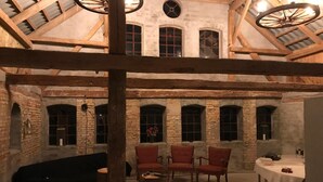 Una habitación rústica con vigas de madera a la vista, paredes de ladrillo y un techo alto con tragaluces. Hay cuatro sillas tapizadas en rojo y una mesa redonda con mantel blanco.