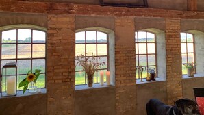 Una habitación rústica con tres ventanas de arco que dan a un campo, un cuadro en la pared y un perro sentado junto a una ventana.