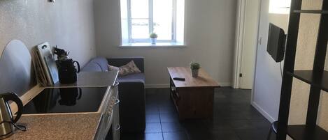 A compact kitchen with a sink, stove, and countertop. There's a window with a view, a small table with a plant, and a dark floor.