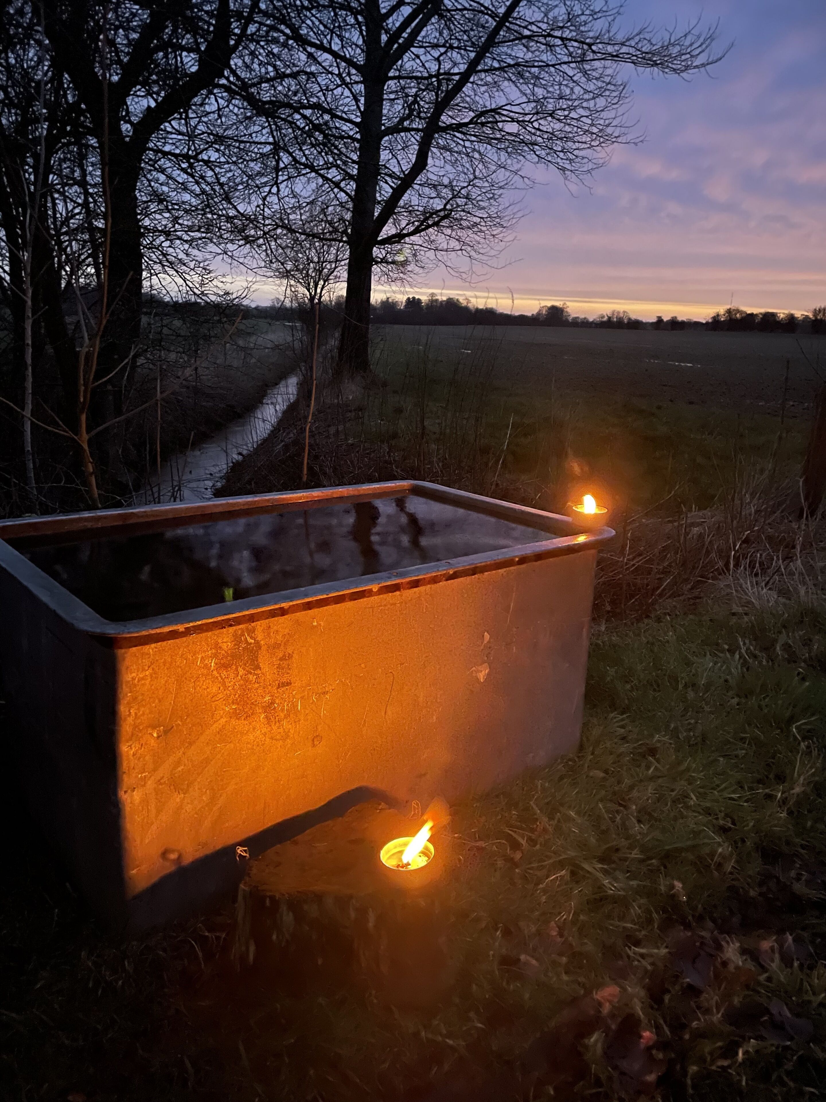 Ein Metallbehälter mit zwei brennenden Kerzen im Freien, mit einem Feld und Bäumen im Hintergrund.