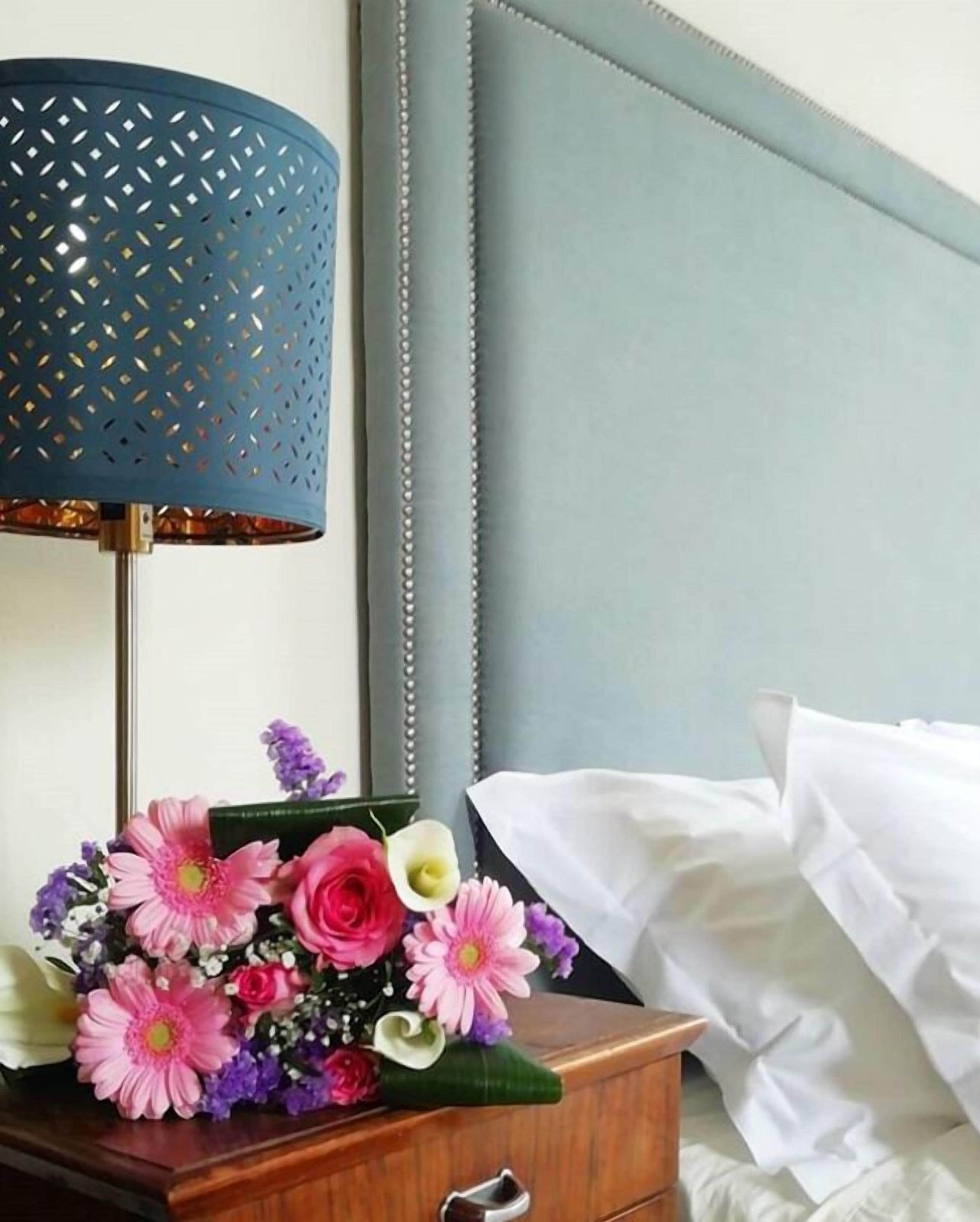 Un lit bien fait, recouvert d’une couette blanche, une table de chevet en bois avec une lampe et un arrangement floral.