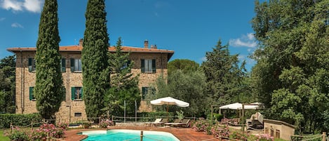 Una villa de piedra con piscina, rodeada de abundante vegetación y altos cipreses.