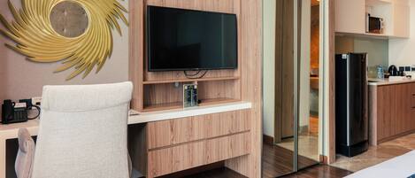 A modern hotel room with a wooden desk, a flat-screen TV, a chair, a microwave, a refrigerator, and a glass partition.