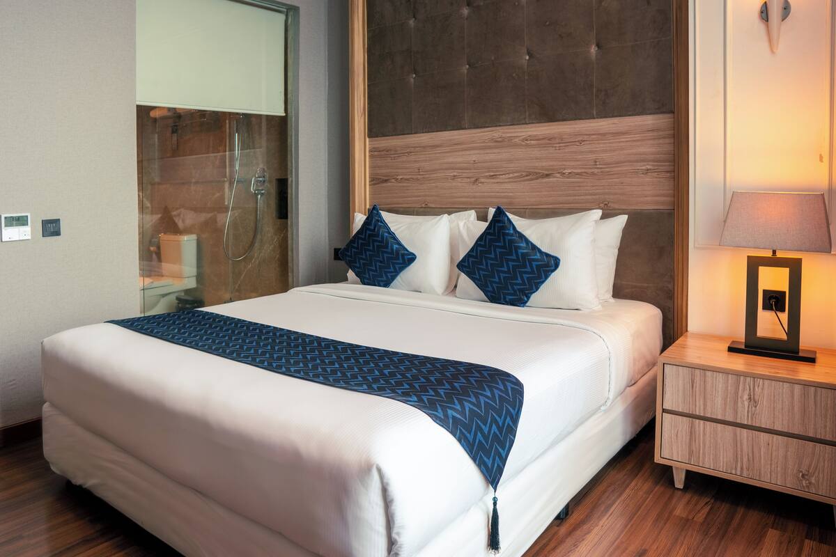 A hotel room with a large bed, a wooden headboard, a nightstand with a lamp, and a glass shower enclosure.