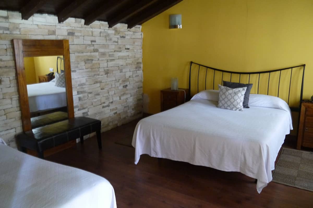 Quarto com parede de pedra, banco de madeira, cama com roupa de cama branca e cabeceira de metal preto.