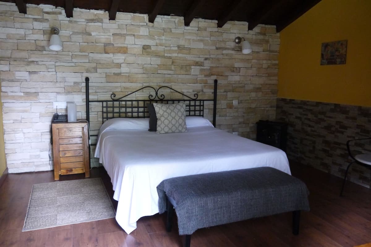 Quarto com parede de pedra, estrutura de cama de madeira, mesa de cabeceira com abajur e um banco.