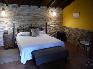 A bedroom with a stone wall, a bed with white bedding, a bench, a chair, and a small table with a lamp.