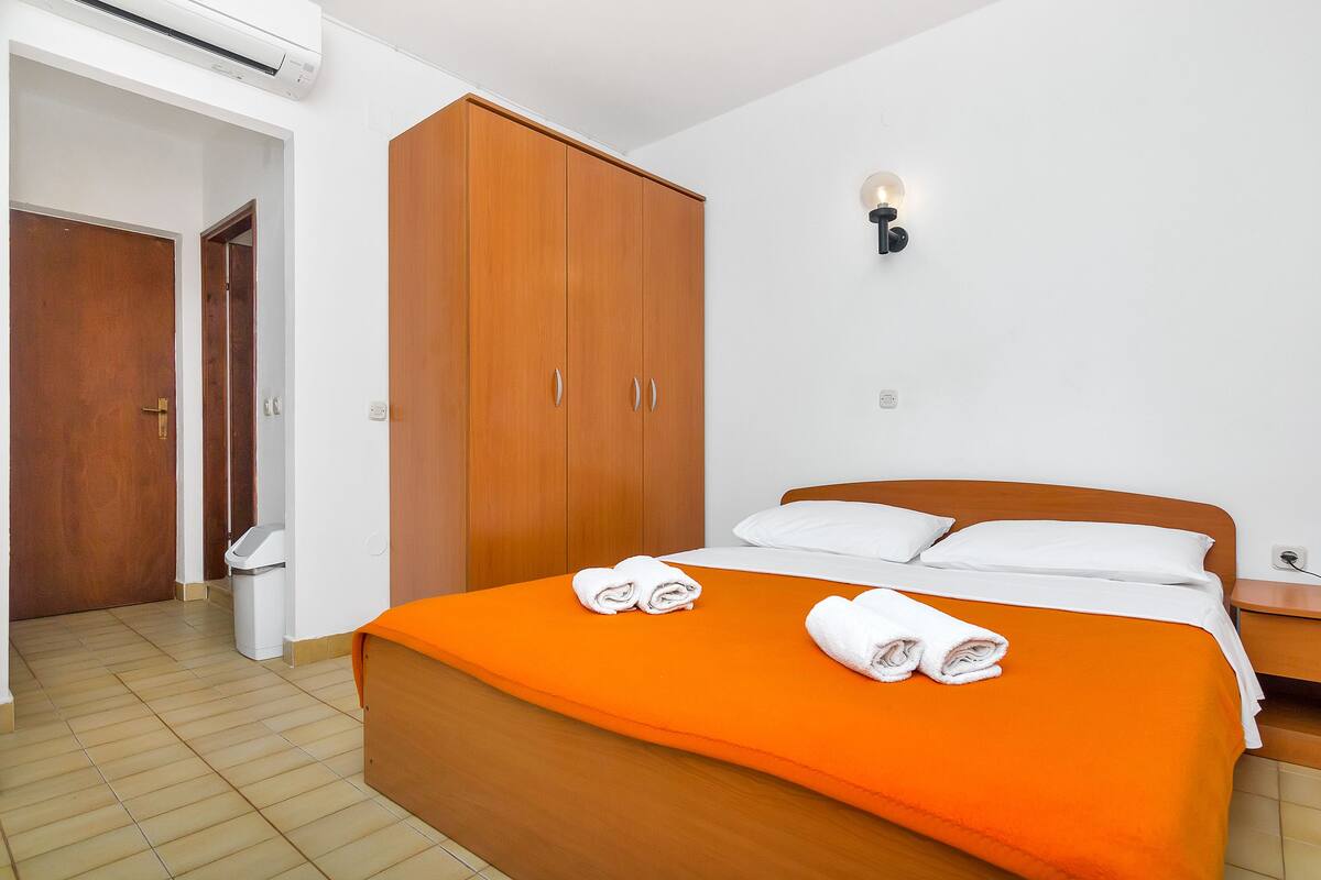 Quarto de hotel com guarda-roupa de madeira, cama com colcha laranja e luminária fixada na parede.
