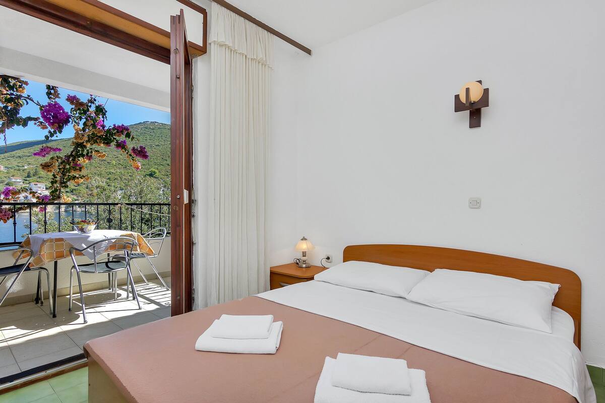 Quarto de hotel com uma cama, uma mesa com cadeiras, uma mesa de cabeceira com abajur e uma varanda com vista para as colinas.
