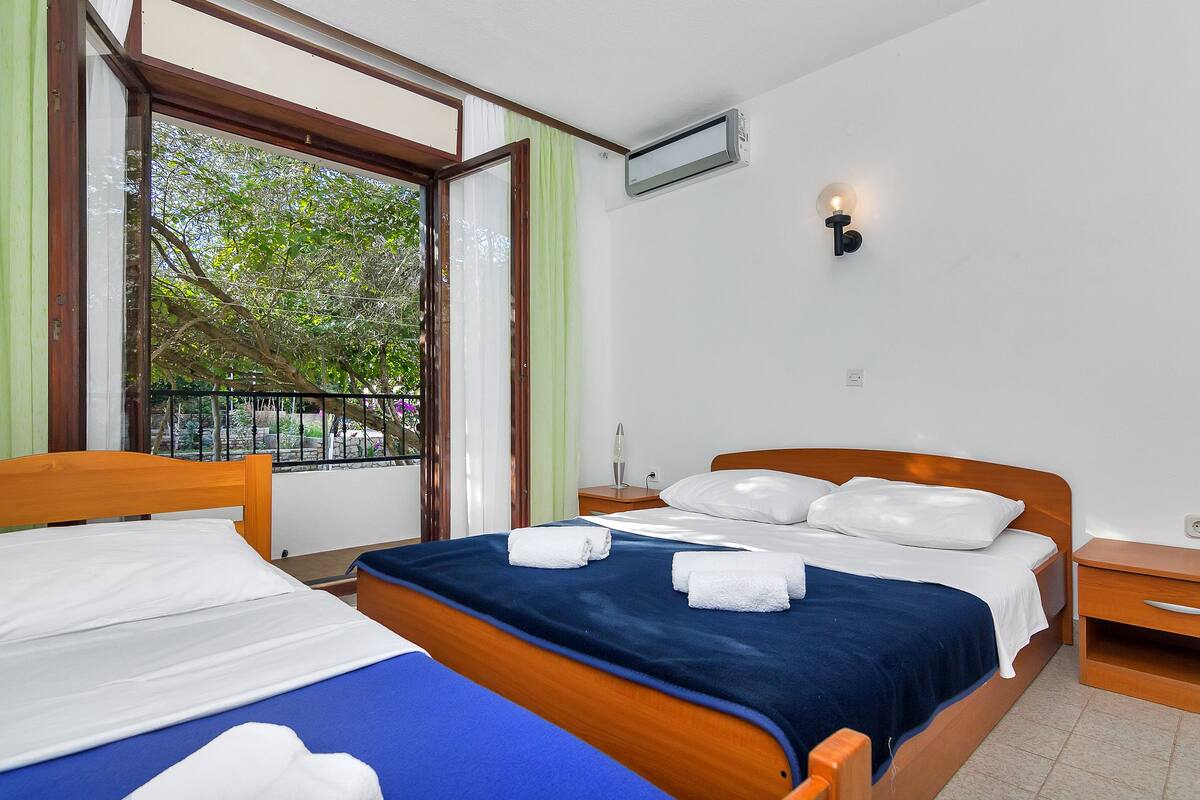 Quarto de hotel com duas camas, uma janela com vista para árvores, ar condicionado fixado na parede e mesa de cabeceira com abajur.