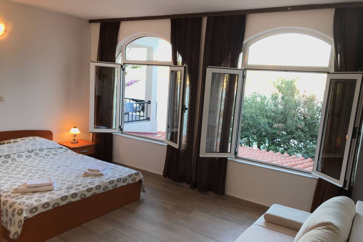 Quarto com cama, mesa de cabeceira, luminária, sofá e janelas amplas com cortinas.