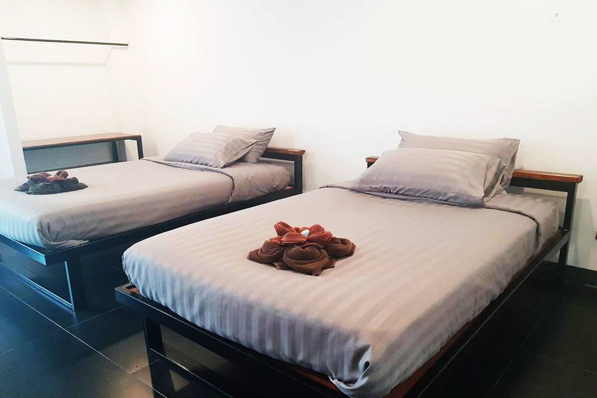 Two single beds with grey linens and wooden frames, a small bedside table with a lamp, and a neatly folded towel on the bed.