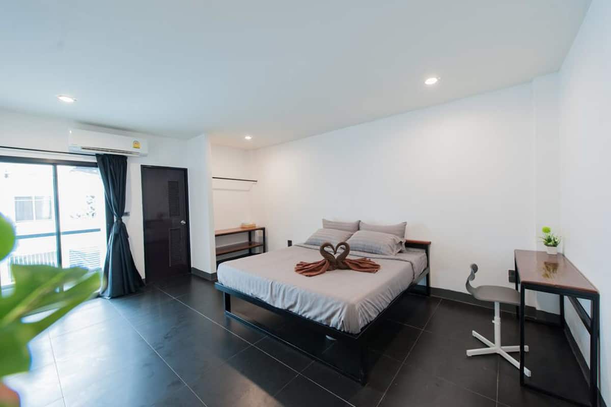A modern hotel room with a large bed, a desk, and a chair. There is a window with curtains, a plant, and an air conditioning unit.