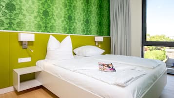 A hotel room with a bed, green wallpaper, white bedding, a magazine on the bed, and a window with a view of greenery.