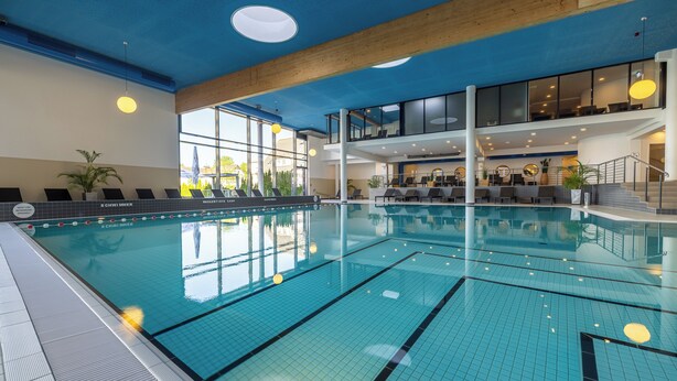 An indoor swimming pool with clear water, a tiled floor, and a modern design with a blue ceiling and large windows. - Carat Residenz-Apartmenthaus (Groemitz)