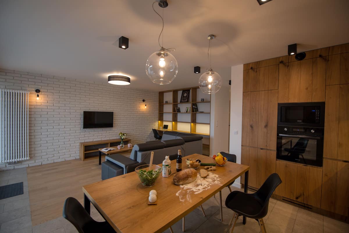 A modern kitchen with a wooden table, chairs, and a dining area. There is a TV mounted on the wall, a microwave, and a wooden cabinet.