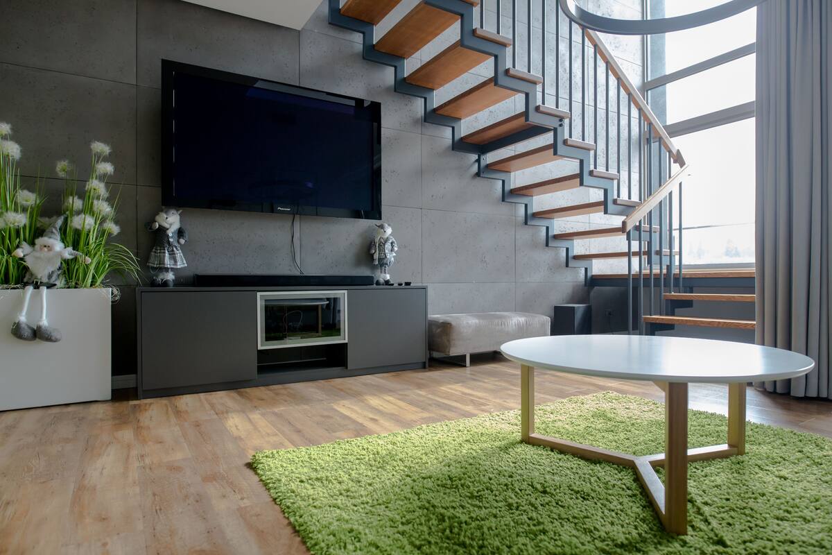 A modern living room with a flat-screen TV, a staircase with wooden steps, a round table, and a green rug.