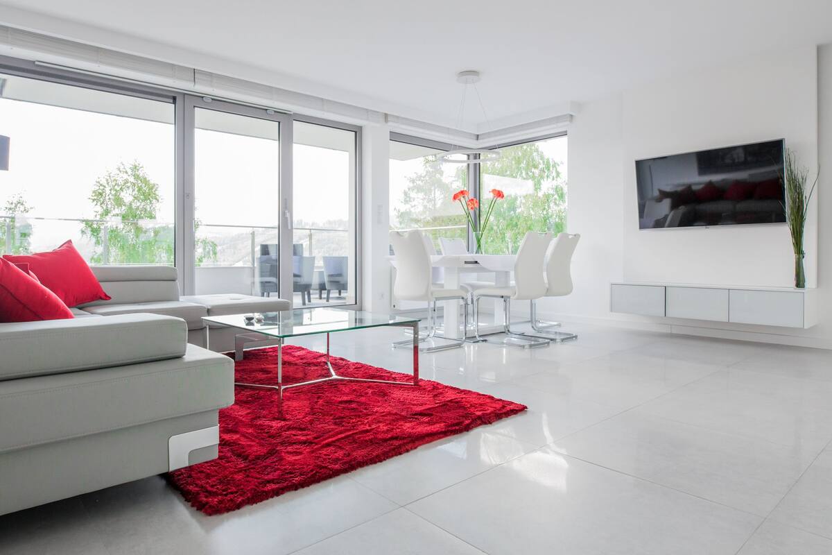 A modern living room with a white sofa, red cushions, a glass coffee table, and a red rug. There is a dining area with white chairs and a table, and a large window offering a view of greenery outside.