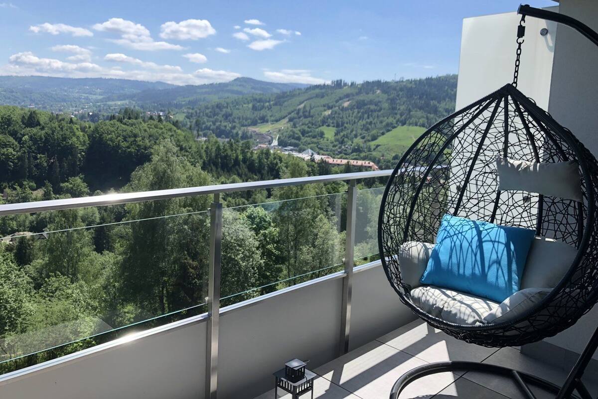 A balcony with a hanging chair, a view of a forested valley, and a clear sky.