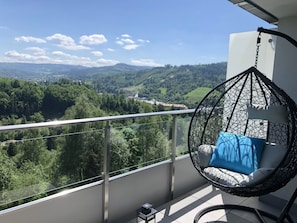 A balcony with a hanging chair, a view of a forested valley, and a clear sky.