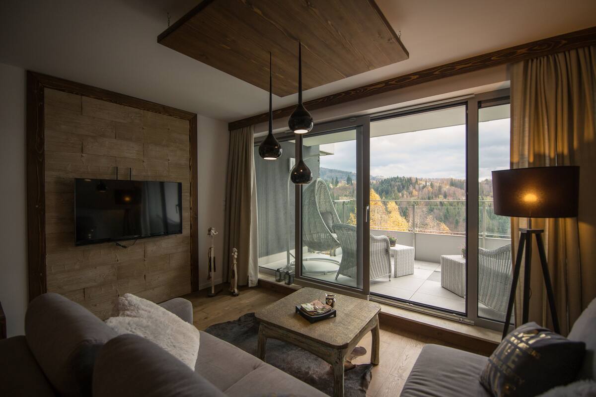 A modern living room with a flat-screen TV mounted on a wooden wall, a sofa, a coffee table, and a large sliding glass door leading to an outdoor balcony with a view of trees and a cloudy sky.