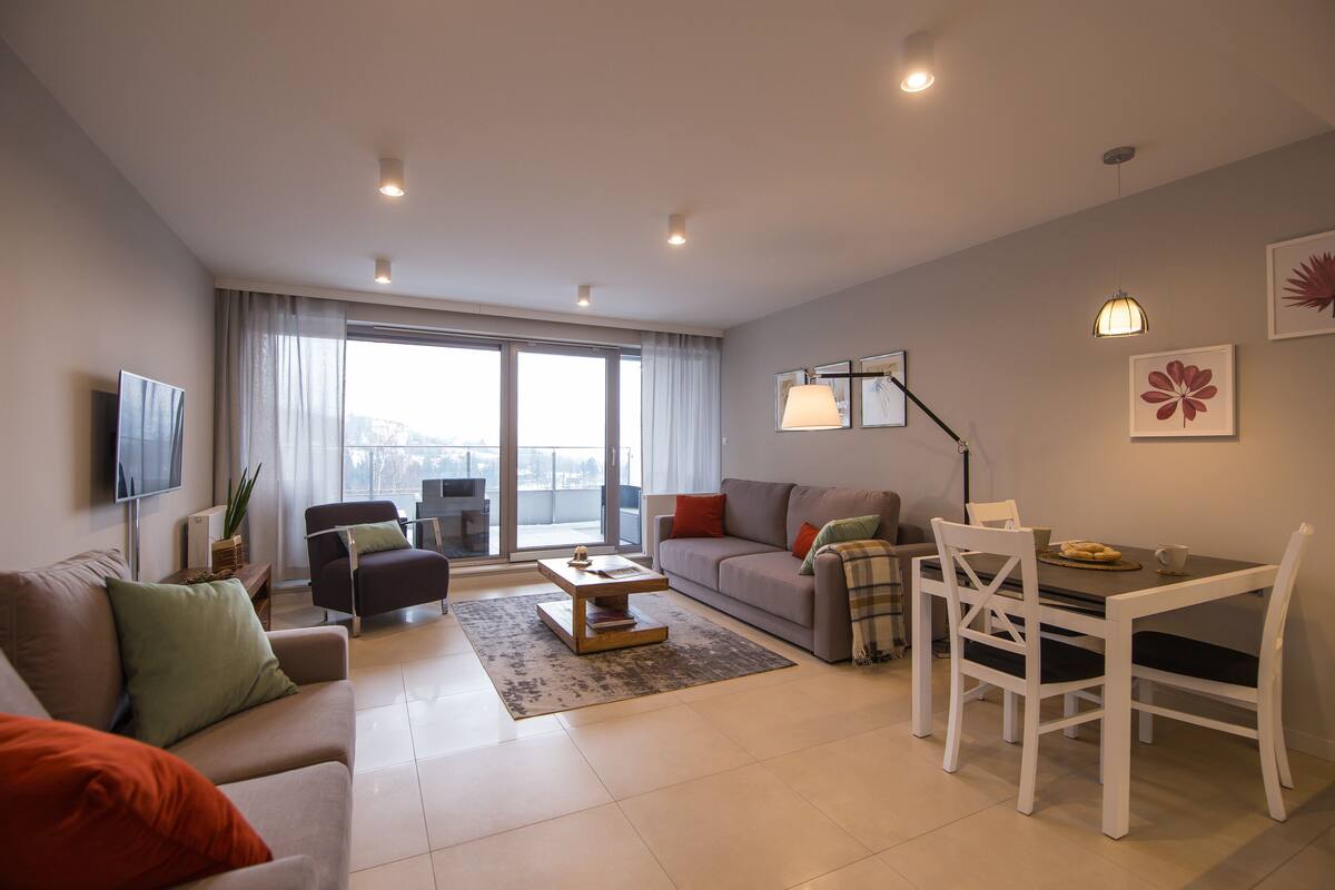 A modern living room with a sofa, coffee table, dining table, and chairs. There is a TV mounted on the wall and a window with curtains.