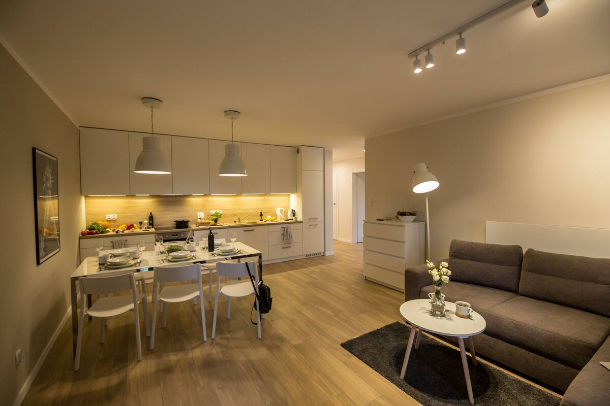 A modern kitchen with white cabinets, a dining table with chairs, and a small side table with a vase and cup.
